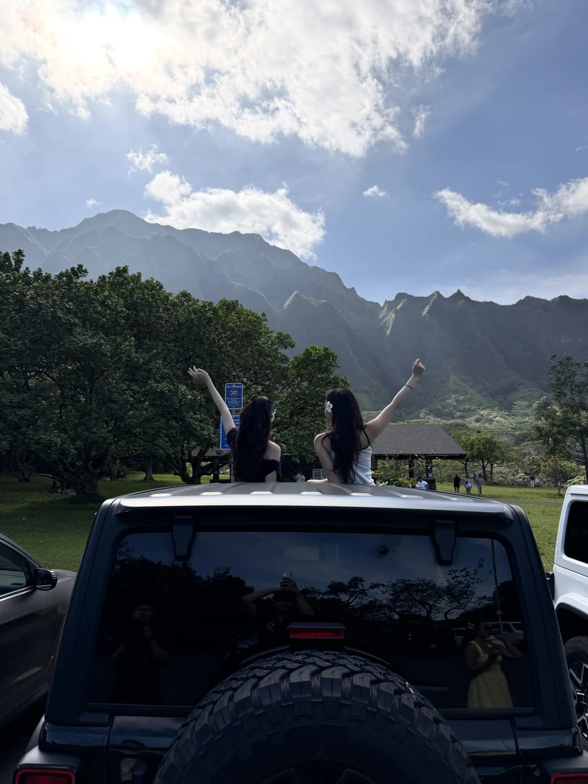 Two friends standing through a vehicle roof beneath green mountains and a blue sky
